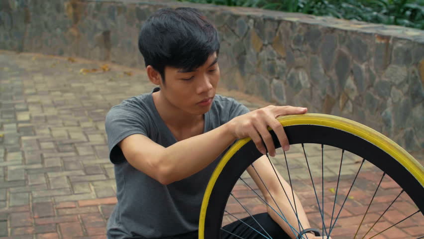 Medium shot of young Asian man sitting on paved road and speeding bike wheel