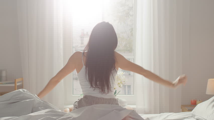 Beautiful Brunette Slowly Waking up in the Morning, Stretches and Gets Up from the Bed, Sun Shines on Her From the Big Window. Happy Young Girl Greets New Day Royalty-Free Stock Footage #1023333379