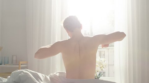 Handsome Fair Boy Sleeping Cozily in Bed, Slowly Wakes up, Gets out of Bed and Stretches Lazily. Young Caucasian Man. Early Morning Sun Shines Through the Window.