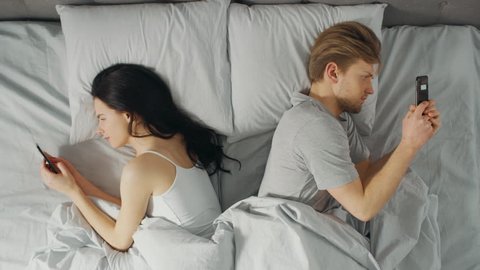 Alienated Millennial Young Couple in the Bed, Young People Turn Away From Each other Using Smartphones, Browsing Through Social Networks and Not Talking to Each Other. Top Down Camera Shot