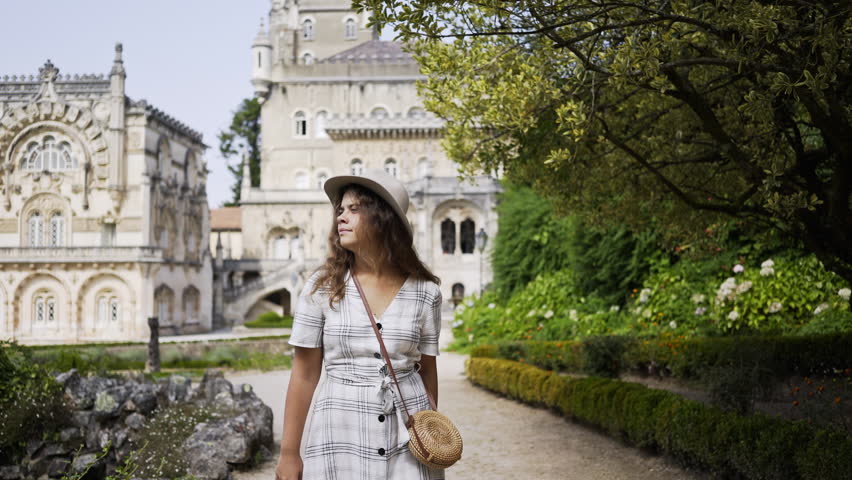 Pretty young woman in hat and dress walking on alley of beautiful park, smiling and looking around at blurred background historic building