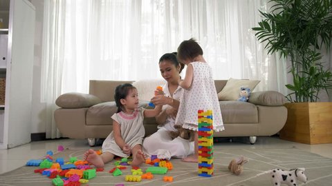Wide shot of young Asian woman sitting on carpet in living room with two little Asian girls and playing with tinker toys