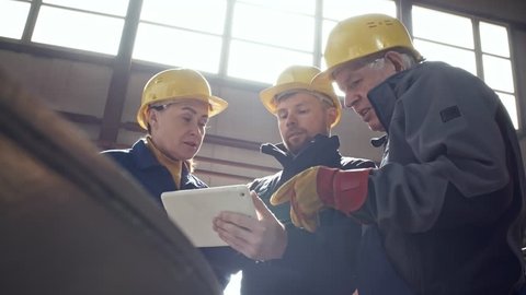 Low angle shot of female and male engineers in gloves and hard hats looking at tablet and discussing project at factory