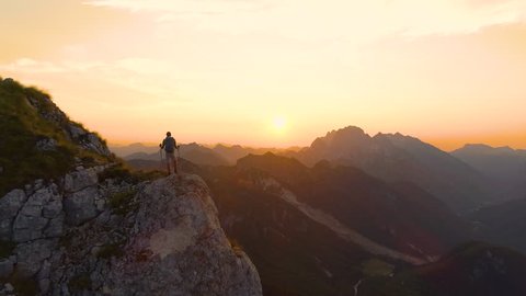 AERIAL, SILHOUETTE: Excited hiker outstretches arms as he watches the sunset from a high cliff in the picturesque Slovenian Alps. Unrecognizable man on active summer vacation watching the sunrise.