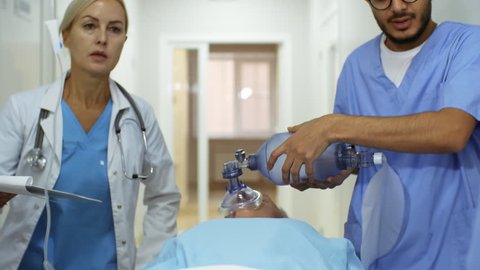 Dolly shot of female ER doctor with clipboard walking and giving instructions to nurses administering oxygen and IV fluids to patient while pushing gurney and rushing him to surgery