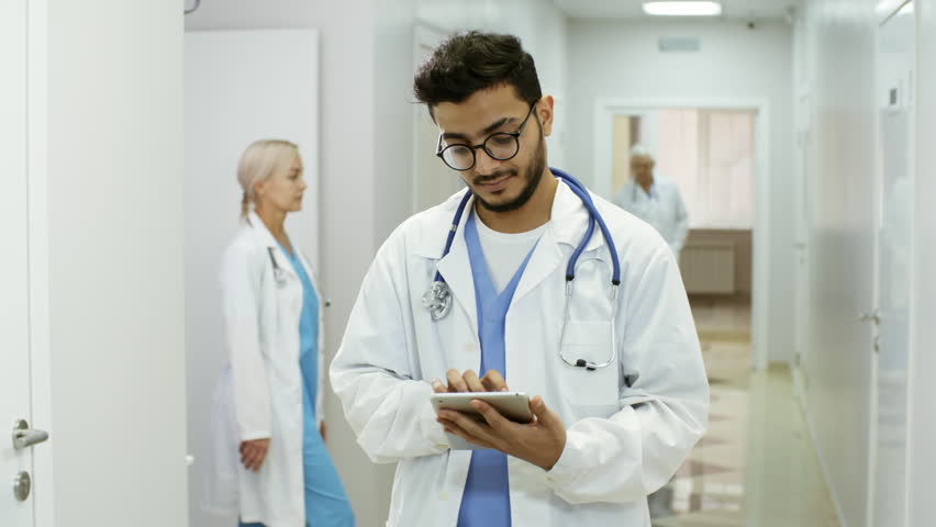 Tracking shot of cheerful Arab male physician in lab coat standing in busy hospital corridor and working on tablet, then looking at camera and smiling