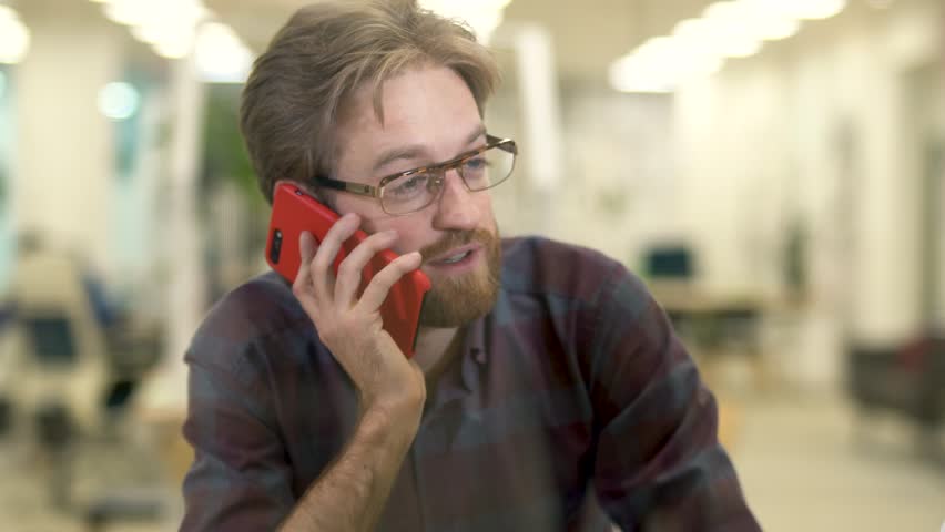 Bearded smiling guy with glasses and casual plaid shirt talking on a cell phone sitting with a girl at the table.