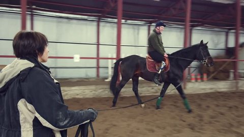 in special hangar, a young disabled man learns to ride a horse with close supervision teacher, hippotherapy. man has artificial limb instead of his right leg. rehabilitation of disabled with animals.