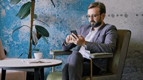 Pleased bearded business man in eyeglasses using smartphone while sitting by the table at cafe