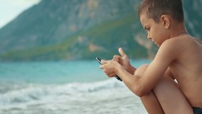 Thoughtful boy using smartphone near sea waves. Child texting messages on mobile phone sitting on seaside. Teenager boy watching video online at phone during summer holidays