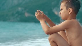 Boy playing games on smartphone while sitting on beach. Boy playing with mobile phone and relaxing at sea. Teen using smartphone sitting on ocean shore during summer holidays