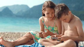 Happy kids play games on mobile phone at sea beach. Sister and brother enjoying summer vacation at ocean with mountains on background. Children playing smartphone game together