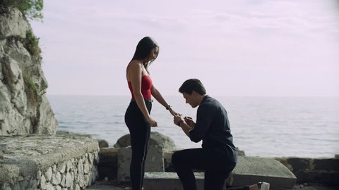 Loving Italian boyfriend proposes to his girlfriend who happily accepts and they hug each other, with view of the ocean and coast line in the background, on bright sunny day. 