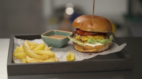 The camera moves bottom up and stops at the shelf with a tray. A burger fixed with a stick, French fries and a sauceboat are lying on the tray