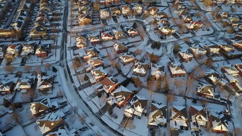 Aerial drone footage of a suburban community in Canada after a blizzard snow storm.  Snow fills the field of view and everything is coated in white the sun sets in the distance.