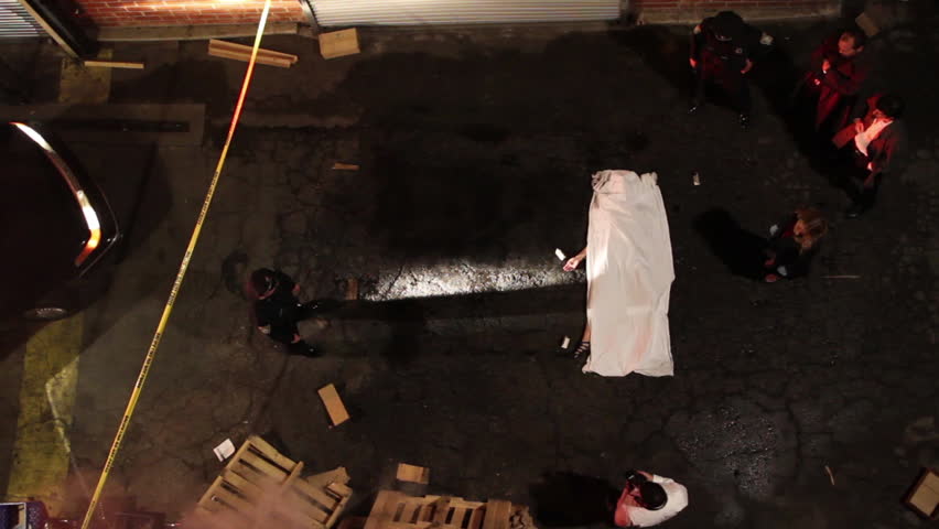Overhead shot of an alley at night with an active homicide crime scene being investigated by law enforcement. Police and detectives walk around the victim's body while a crime scene photographer takes