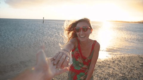 Girl leading boyfriend to the beach at sunset. Slow motion shot of woman holding lover's hands on the beach and leads him towards the sunset. Follow me concept point of view 