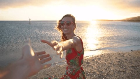 Girl leading boyfriend to the beach at sunset. Slow motion shot of woman holding lover's hands on the beach and leads him towards the sunset. Follow me concept point of view 