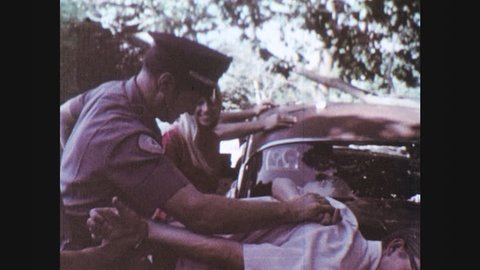 1960s: police officers force teenage boy in eyeglasses onto trunk of car, struggle, restrain and slap handcuffs on wrist and drag away man past ambulance and patrol cruiser in grass parking lot.