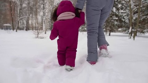 woman is holding hand of her infant baby and learning it to go in winter day, stepping over snow