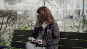 Concentrated curly woman typing on tablet while sitting on wooden bench. Focused brunette using tablet in park. Technology concept
