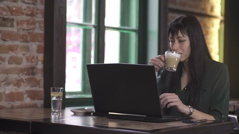 Young business woman ending conversation on cell phone, she sitting at table and starts typing on laptop and drinking latte coffee, girl working in coffee shop, slide shot from lady, dolly, daylight.