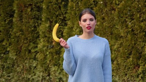 Portrait attractive young woman in blue jacket holds banana in her hand then lowers it and lifts it up imitating male erection, talking oops and smiling against green trees