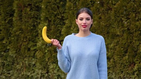 Portrait attractive young woman in blue jacket holds banana in her hand then lowers it and lifts it up imitating male erection, talking oops and smiling against green trees