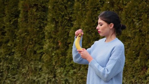 Portrait seductive attractive young woman with dark hair and red lips in blue jacket peeling banana and biting it, then she looks straight to camera on background green trees