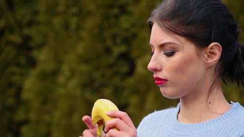 Close-up seductive attractive young woman with dark hair and red lips in blue jacket peeling banana and biting it on blurred background
