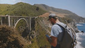 Happy middle aged blogger tourist man with backpack takes panoramic smartphone video of iconic Bixby Creek bridge USA.