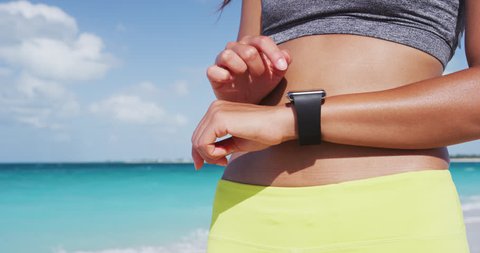 Smartwatch - Sporty woman using smartwatch during exercise. Female runner is standing at beach in summer. She is tracking her fitness after jogging against sea.