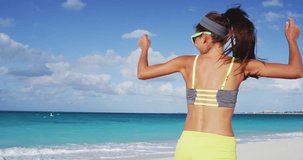 Cheerful sporty woman running dancing laughing having fun on sea shore against sky. Young female in sportswear is wearing sunglasses. Happy brunette is enjoying at beach during summer.