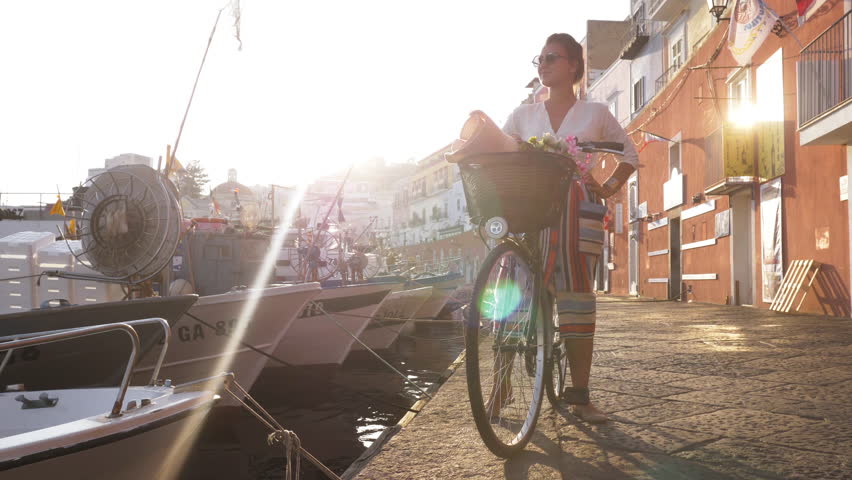 Young woman with vintage bike standing at seafront by the harbor in Ponza island Italy. Fashion colorful dress skirt and hat.