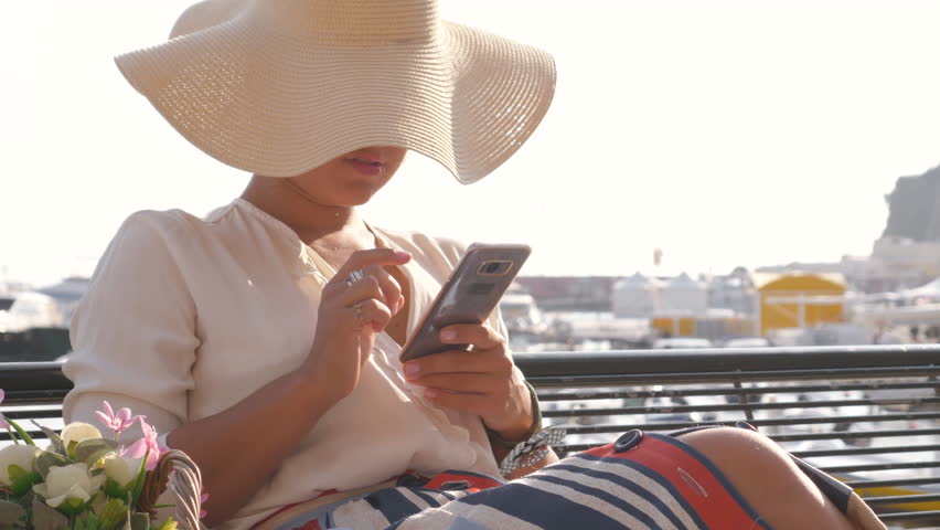 Young woman using smartphone sitting on a bench in Ponza town island, Italy. Fashion colorful dress skirt, large hat, flower basket . Cityscape and sea in background.