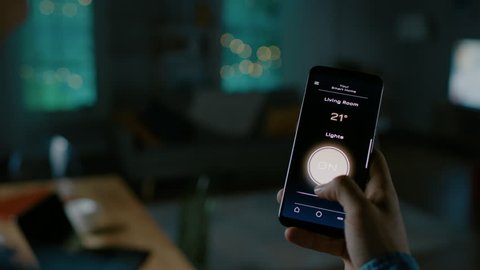 Close Up Shot of a Smartphone with Active Smart Home Application. Person is Tapping the Screen and Light is Being Turned On in the Room. It's Cozy Evening in the Apartment.