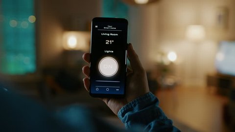 Close Up Shot of a Smartphone with Active Smart Home Application. Person is Giving a Voice Command and Light Turns On in the Room. It's Cozy Evening in the Apartment.