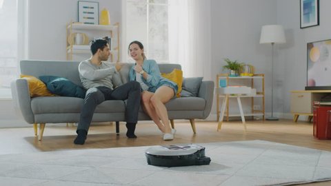 Smart Robot Vacuum Cleaner Sucking Up Dust from a Carpet. Beautiful Couple is Sitting on a Sofa and Talking in the Background. Technological Home Appliance Device Moves Past Them.