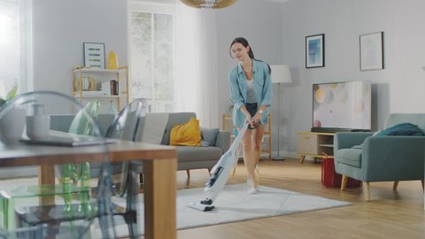 Young Beautiful Woman in Jeans Shirt and Shorts is Vacuum Cleaning a Carpet in a Bright Cozy Room at Home. She Uses a Modern Cordless Vacuum. She's Happy and Cheerful.