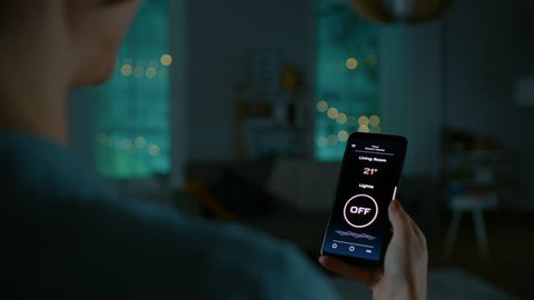 Young Beautiful Woman Gives a Voice Command to a Smart Home Application on Her Smartphone and Lights in the Room are Being Turned On. She Walks and Sits on a Couch. It's a Cozy Evening.