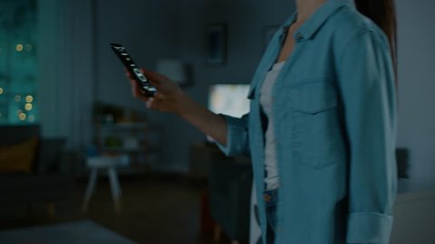Young Beautiful Woman Gives a Voice Command to a Smart Home Application on Her Smartphone and Lights in the Room are Being Turned On. She Walks and Sits on a Couch. It's a Cozy Evening.
