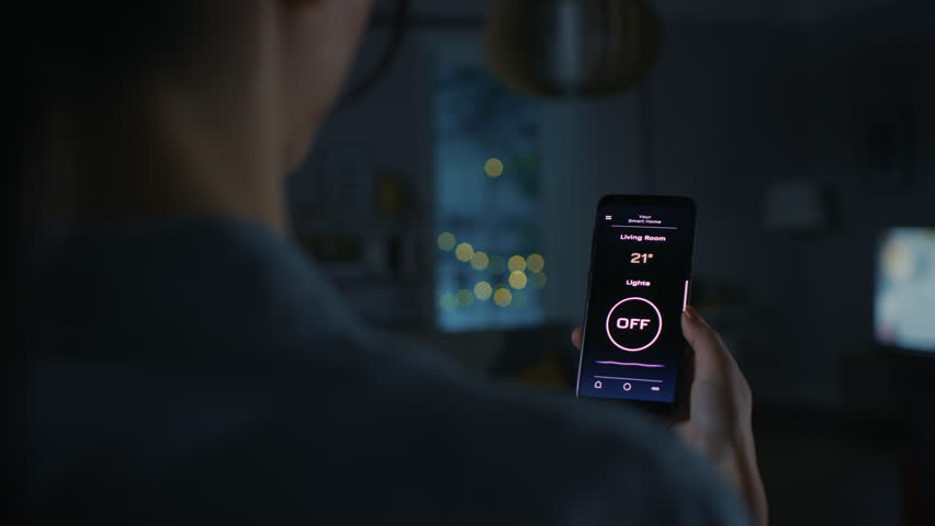 Young Beautiful Woman Gives a Voice Command to a Smart Home Application on Her Smartphone and Lights in the Room are Being Turned On. She Walks and Sits on a Couch. It's a Cozy Evening. | Shutterstock HD Video #1023495835