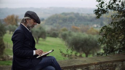 Wide shot on 8k helium RED camera. Content Italian man sitting on a stone wall, writing in his book and looking out at the beautiful scenic landscape in Tuscany, on soft overcast day.の動画素材