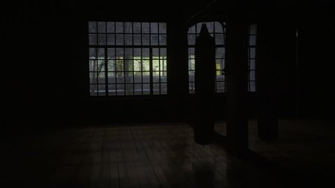 Panning shot from the inside of a boxing gym, filmed behind punching bags. Punching bag on the background of a window in a dark room.