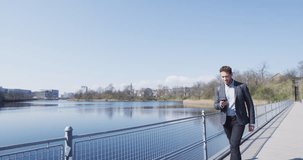 Smiling businessman using smartphone walking on bridge. Confident young male professional is carrying backpack walking by lake in city park. RED Cinema camera in SLOW MOTION.