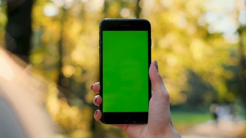 Female hand holding smartphone with green screen. Girl using mobile phone while walking in the autumn park. Back view shot. Chroma key, close up woman hand holding phone with vertical green screen.
