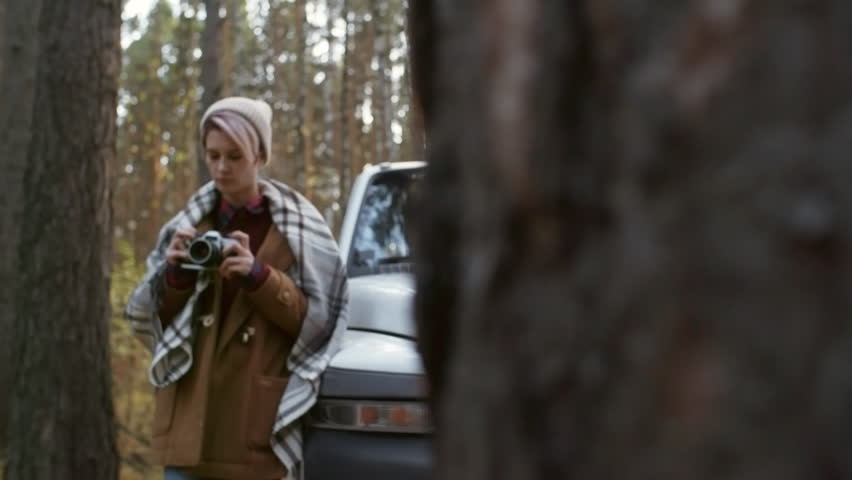 Medium shot of young woman in beanie taking photo with camera during forest trip in autumn