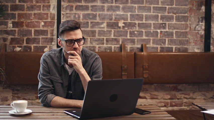 Pensive brunette man in eyewear wearing gray shirt looking away and thinking about something while sitting at the table with laptop in comfortable cafe | Shutterstock HD Video #1023553987