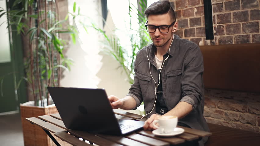 Serious young man wearing earphones checking information on his laptop while sitting at the table in cafe interior | Shutterstock HD Video #1023554014