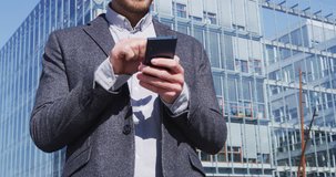 Businessman text messaging on smartphone. Male executive business man on phone is standing by business buildings wearing suit on sunny day.